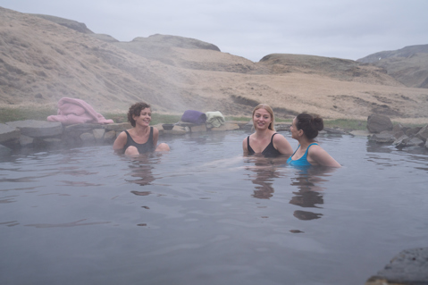 Three women smiling at each other while in a hot spring bath surrounded by mountains