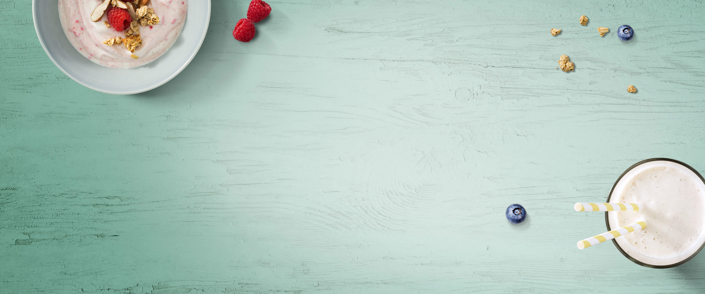 A bowl of yogurt, raspberries and granola and a milk shake against a green wooden backdrop