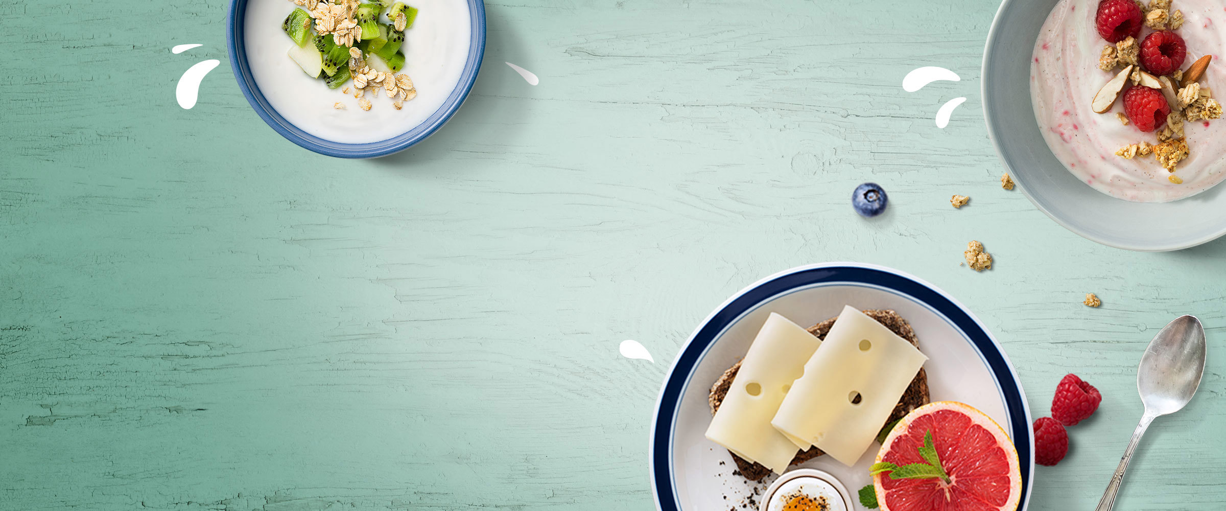 A bowl of yogurt, raspberries and granola, a bowl of yogurt oats and kiwi fruit and a plate of cheese of rye bread and grapefruit against a green wooden backdrop