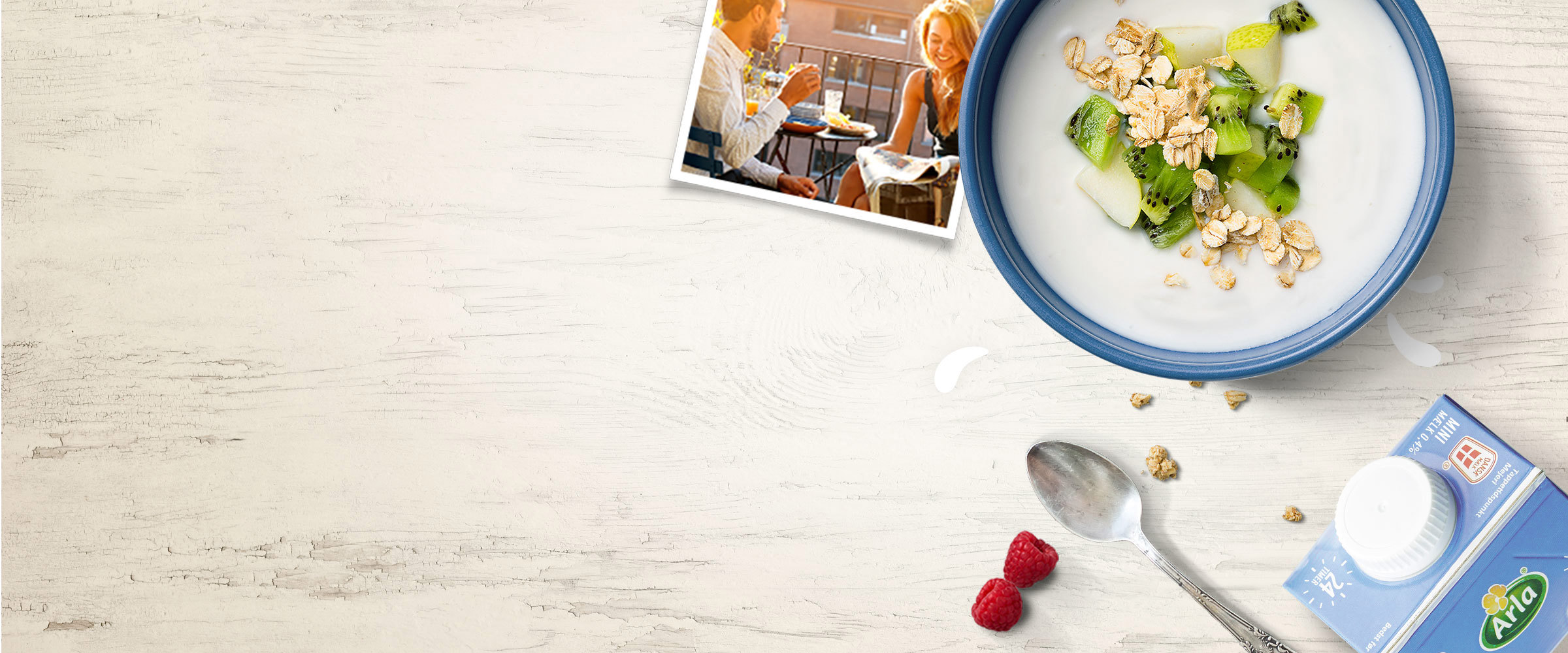 A bowl of yogurt, oats and kiwi fruit, a polaroid of a couple having breakfast, a spoon and carton of arla milk against a cream wooden backdrop