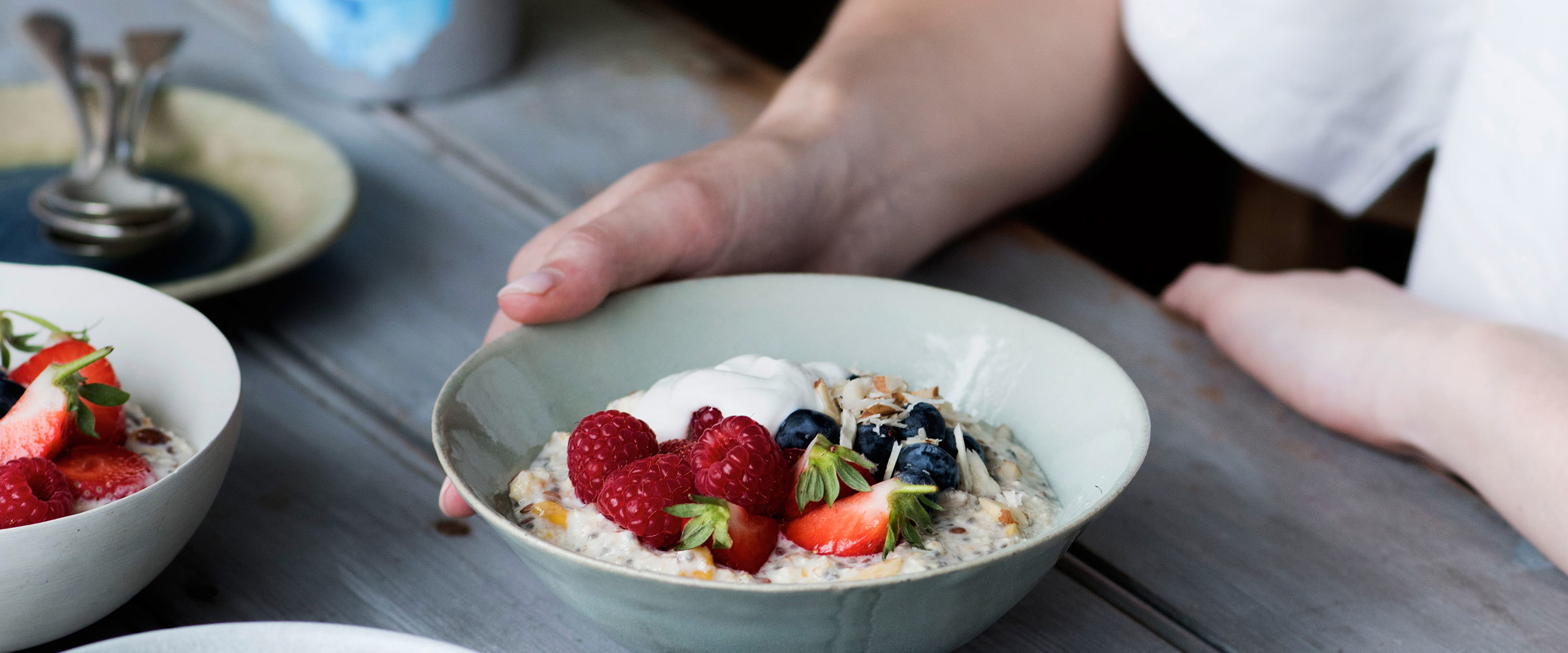 A bowl of bircher muesli topped with berries and yogurt on a table in front of a diners hand
