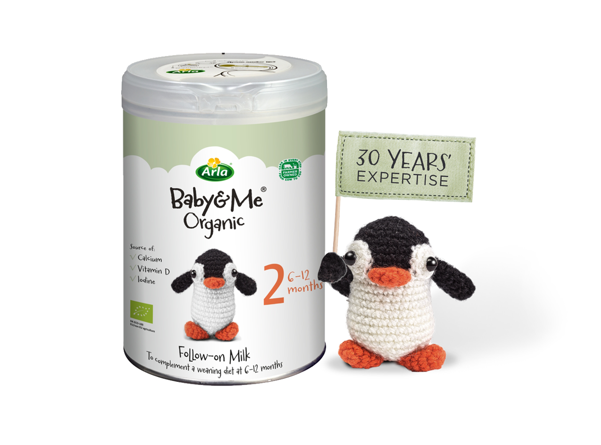 Arla Baby & Me Organic pack next to a knitted penguin toy holding a flag that says 30 years' expertise