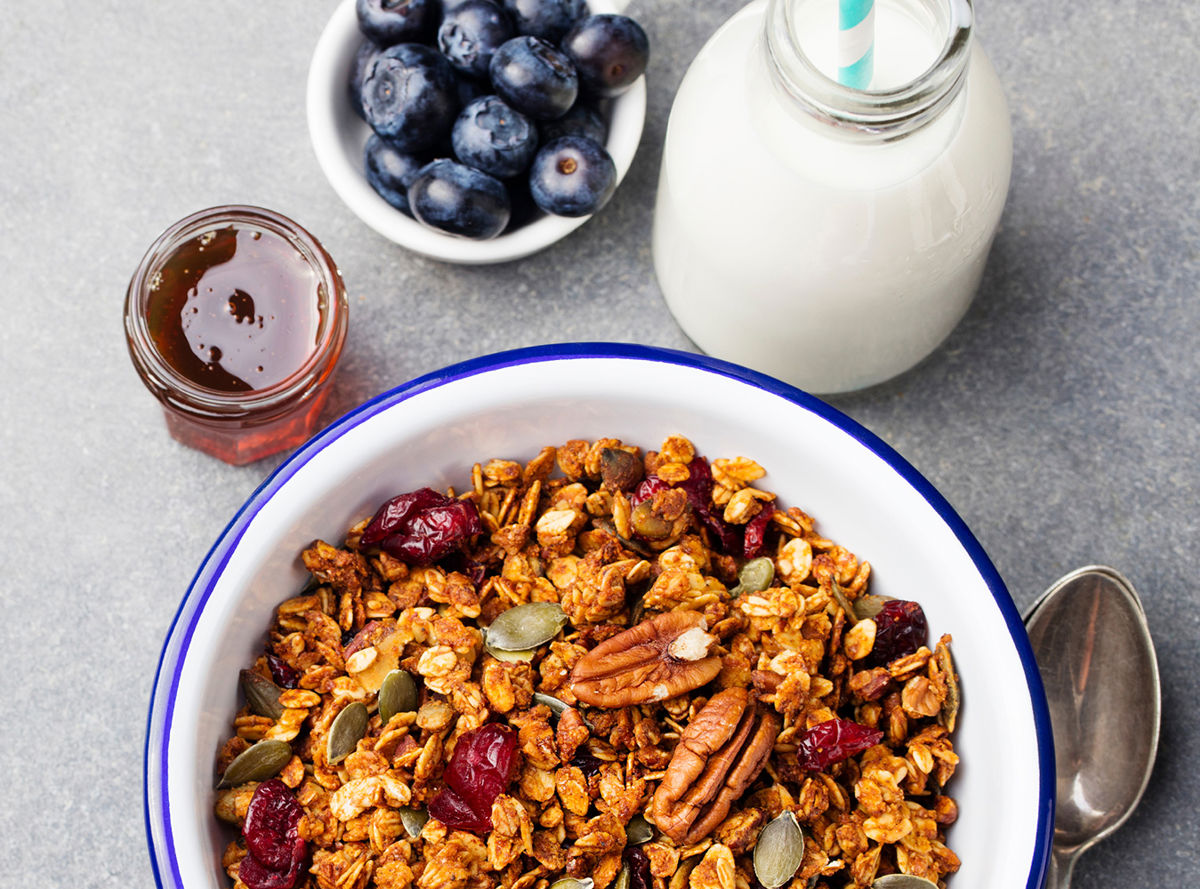 A bowl of oats with nuts, seeds and dried fruit next to blueberries, honey and milk