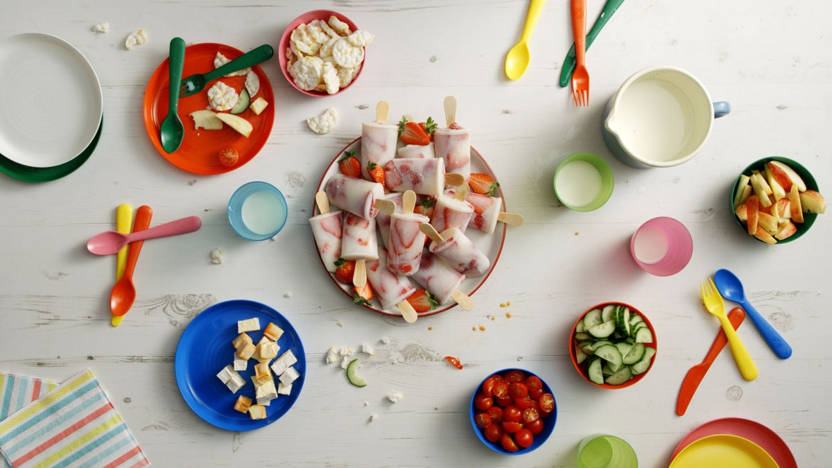 Strawberry ice lollies on a plate surrounded by bowls, cups, and cutlery