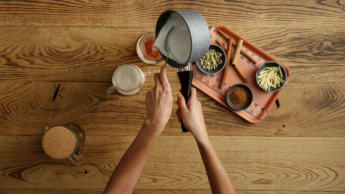 Hot milk being poured from a pot over two glass mugs of chai latte